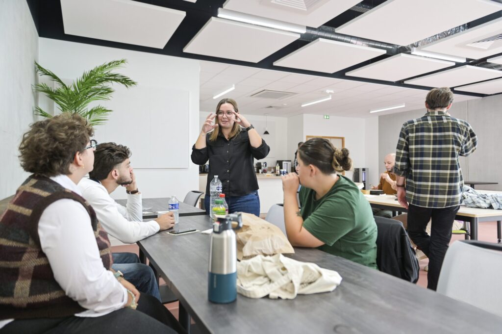 Une intervenante debout explique à cinq participants assis autour d’une grande table de travail