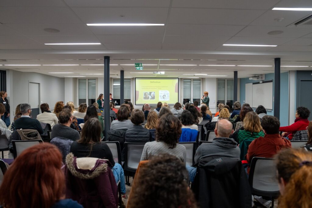 Une salle comble, vue depuis le fond, écoutant une intervenante devant un écran de projection