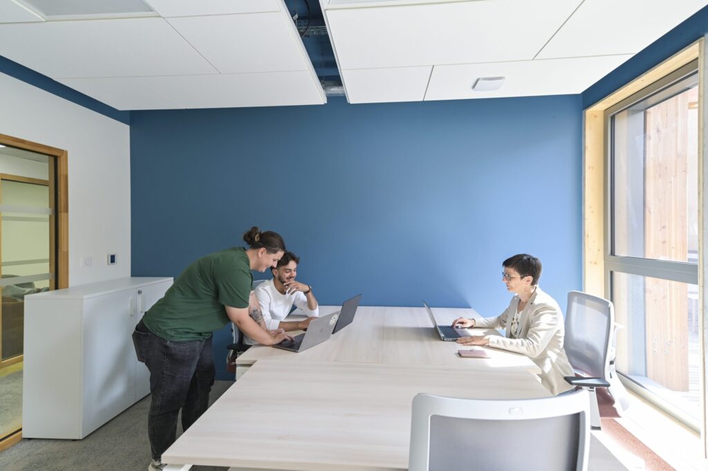 Trois personnes au travail dans un bureau de Niort Tech, grande table claire et mur bleu, baie vitrée sur le patio