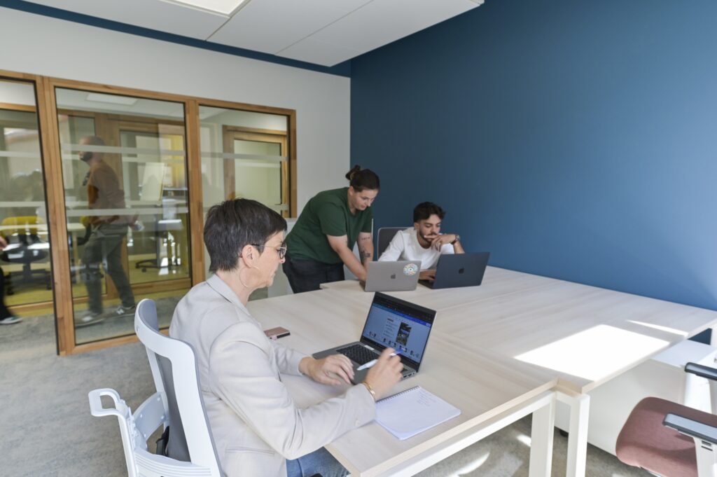 Trois personnes au travail dans un bureau au mur bleu, ouvert sur le couloir vitré