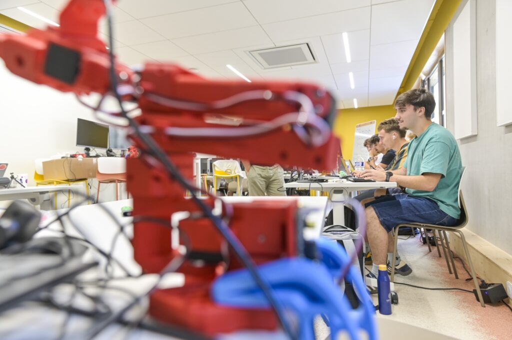 Un bras robotisé au premier plan, des étudiants au travail derrière