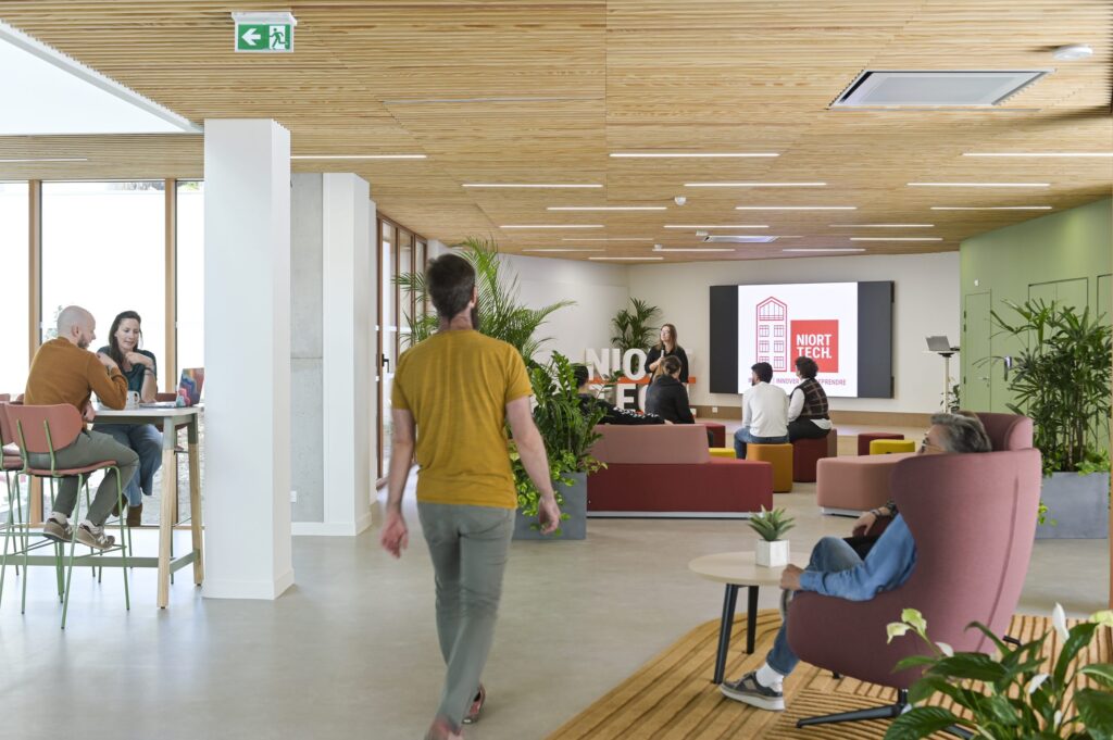 Le hall de Niort Tech : des personnes en réunion informelle, un écran d’accueil et des assises colorées sous un plafond de bois