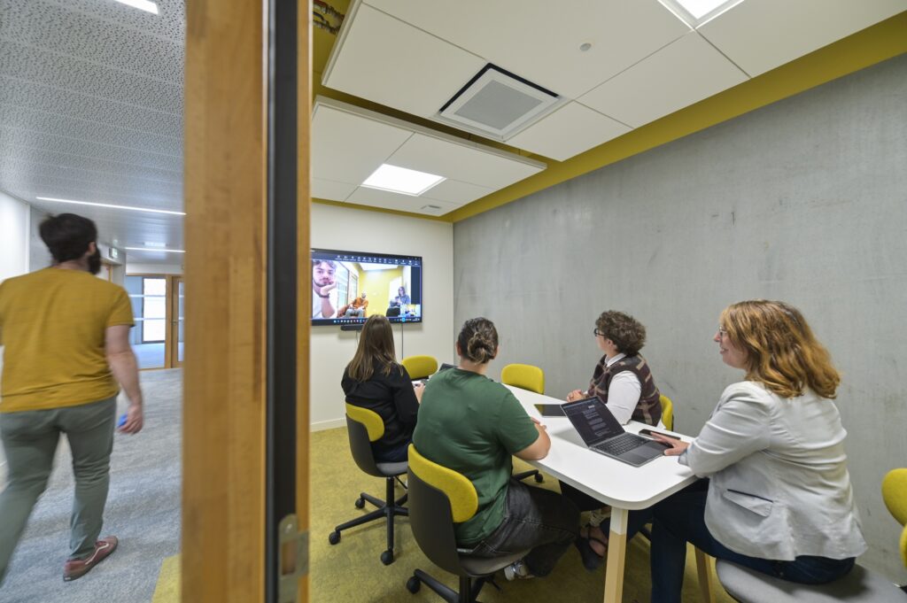 Une réunion dans une salle vitrée, autour d’un écran de visioconférence
